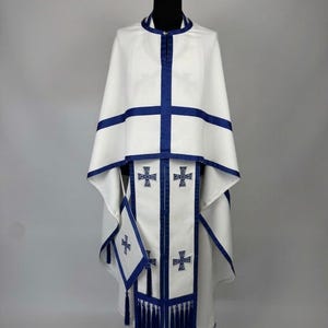 May include: White religious vestment with blue trim and cross details. The set includes a cape, stole, and matching accessories. The design features blue crosses and tassels, creating a formal and ceremonial appearance.