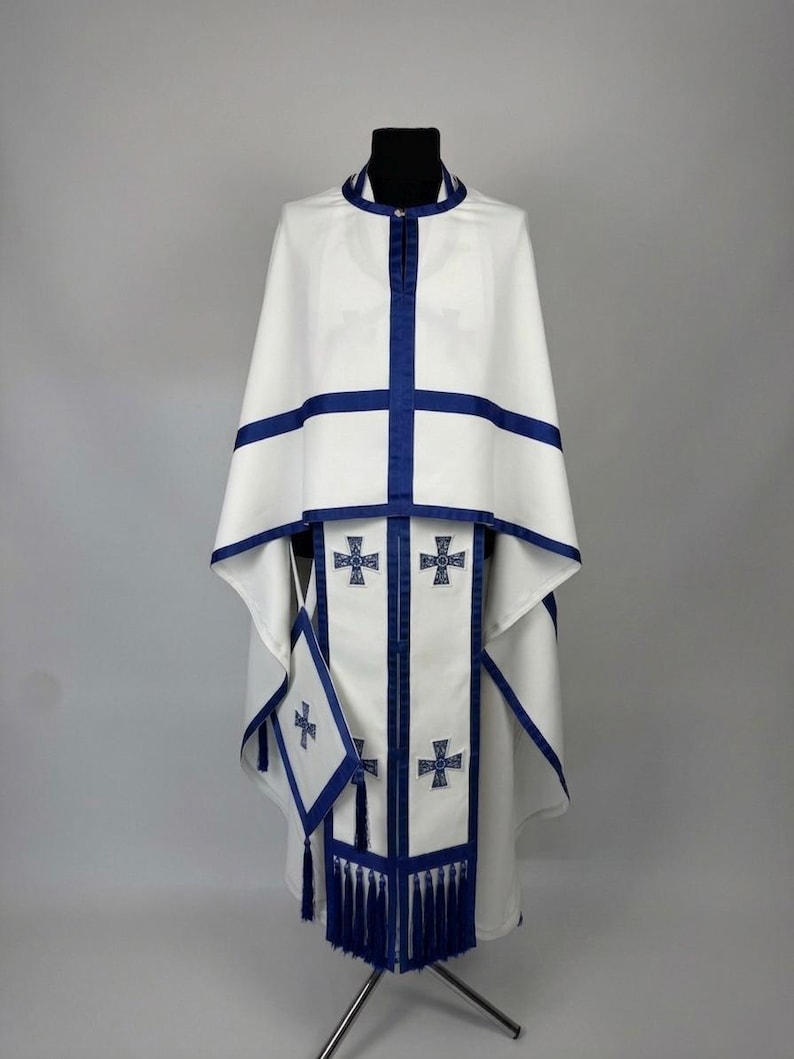May include: White religious vestment with blue trim and cross details. The set includes a cape, stole, and matching accessories. The design features blue crosses and tassels, creating a formal and ceremonial appearance.