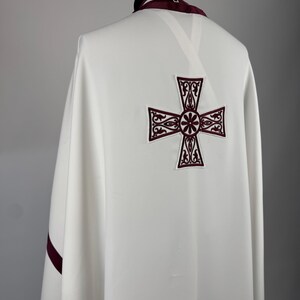 May include: A white liturgical vestment with a large, embroidered burgundy cross on the back. The collar and edges are trimmed with burgundy fabric. The garment is draped over a form, showcasing its design and details.