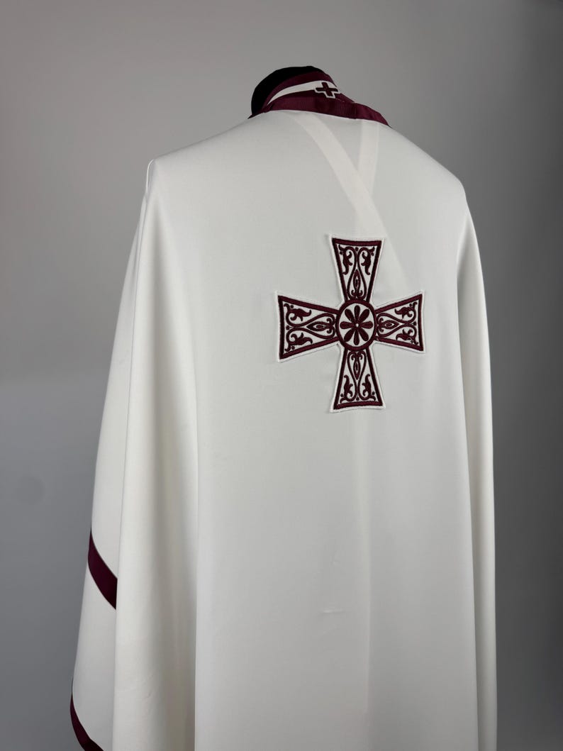 May include: A white liturgical vestment with a large, embroidered burgundy cross on the back. The collar and edges are trimmed with burgundy fabric. The garment is draped over a form, showcasing its design and details.