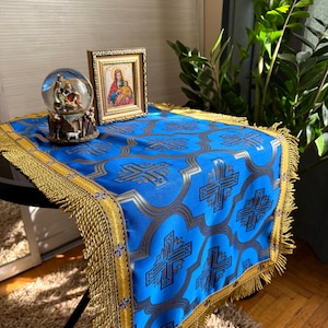 May include: A bright blue rectangular table runner featuring a repeating gold and dark blue cross pattern, finished with a wide gold fringe border. A glass snow globe depicting a nativity scene and a framed icon of the Virgin Mary sit on top.
