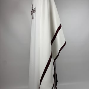 May include: White liturgical vestment with a burgundy cross design on the back and burgundy trim. The garment is draped, showcasing its flowing fabric and detailed embroidery. The vestment is likely used for religious ceremonies.