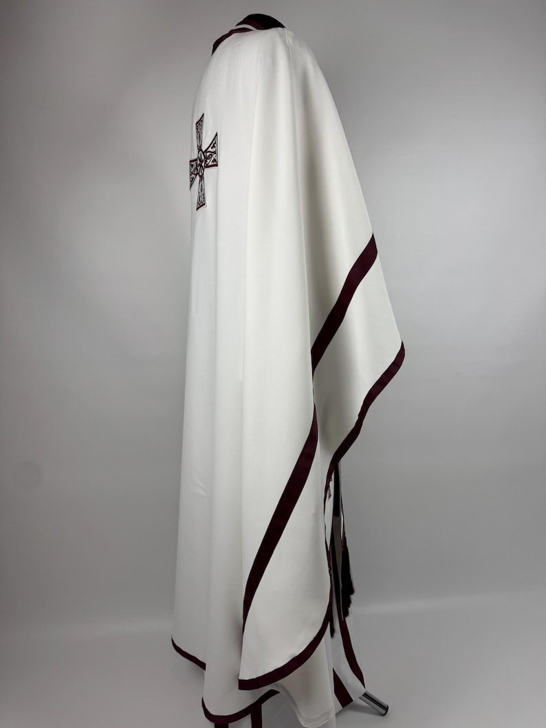 May include: White liturgical vestment with a burgundy cross design on the back and burgundy trim. The garment is draped, showcasing its flowing fabric and detailed embroidery. The vestment is likely used for religious ceremonies.