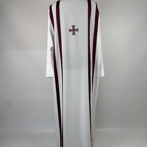 May include: White liturgical vestment with burgundy vertical stripes and a cross emblem on the back. The long-sleeved garment is displayed on a black mannequin head against a plain background. The fabric drapes to the floor.