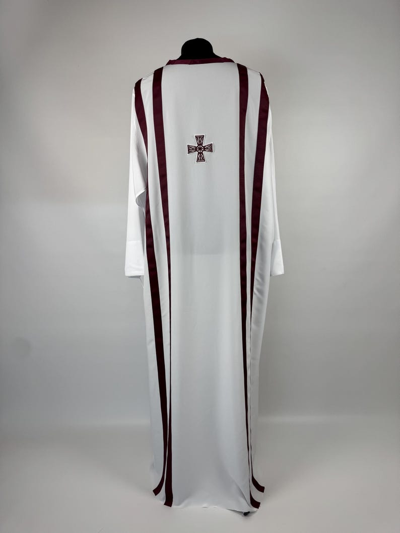 May include: White liturgical vestment with burgundy vertical stripes and a cross emblem on the back. The long-sleeved garment is displayed on a black mannequin head against a plain background. The fabric drapes to the floor.