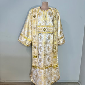 Op de afbeelding: Een wit en goud liturgisch gewaad bestaande uit een tuniek met lange mouwen en een bijpassende stola met ingewikkelde geweven patronen. De stof toont herhalende kruismotieven in gouddraad op een witte achtergrond met gouden randen.