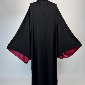 May include: A long, black robe with wide sleeves and a mandarin collar. The interior of the sleeves is lined with a deep red satin fabric. The robe has a button closure at the neck and a flowing silhouette, suitable for formal occasions or costume use.
