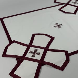 May include: White cloth with a burgundy border and cross-shaped accents. The design features a large cross shape at the bottom and smaller crosses throughout. The fabric appears to be a smooth, possibly linen or cotton material.