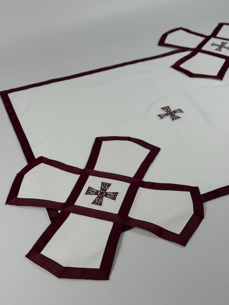 May include: White cloth with a burgundy border and cross-shaped accents. The design features a large cross shape at the bottom and smaller crosses throughout. The fabric appears to be a smooth, possibly linen or cotton material.