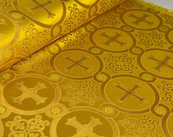 May include: A roll of golden yellow fabric featuring a repeating pattern of embroidered crosses enclosed in circular frames, interspersed with intricate floral scrollwork motifs. The textile has a smooth, satin-like finish with a reflective sheen.