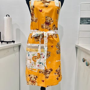 May include: An orange apron featuring a repeating pattern of teddy bears with red bows. The apron has white trim, a patterned pocket, and a matching tie. The straps are white with a colourful pattern.