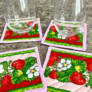 May include: Four square fabric coasters with a strawberry design. Each coaster features a pink border and a quilted pattern. The strawberry pattern includes red strawberries, green leaves, and white flowers. Two clear stemless wine glasses rest on two of the coasters.