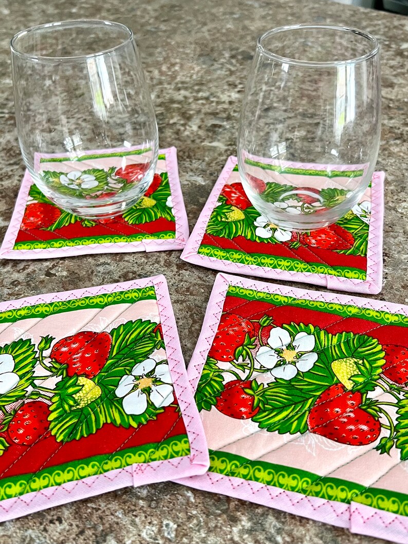 May include: Four square fabric coasters with a strawberry design. Each coaster features a pink border and a quilted pattern. The strawberry pattern includes red strawberries, green leaves, and white flowers. Two clear stemless wine glasses rest on two of the coasters.