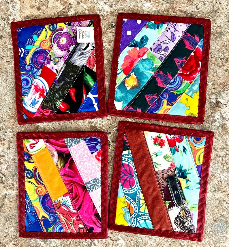 May include: Set of four handmade fabric coasters. Each coaster features a patchwork design with diagonal strips of colorful patterned fabric, including floral, abstract, and geometric prints. The coasters are bordered with a dark red fabric and have a square shape.