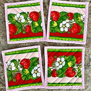 May include: Set of four square fabric coasters featuring a strawberry design. Each coaster has a pink border with a decorative stitch, and a quilted pattern. The strawberry design includes red strawberries, green leaves, and white flowers.