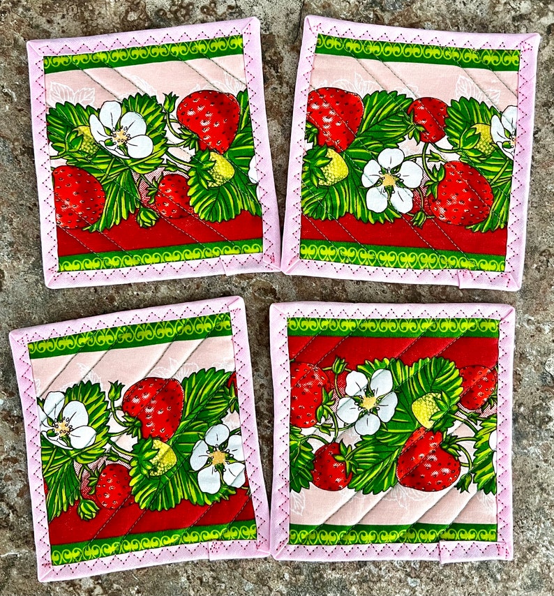 May include: Set of four square fabric coasters featuring a strawberry design. Each coaster has a pink border with a decorative stitch, and a quilted pattern. The strawberry design includes red strawberries, green leaves, and white flowers.