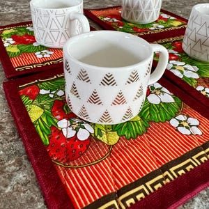 May include: White ceramic mugs with geometric patterns rest on red and green strawberry-themed placemats. The mugs have handles and are decorated with gold and black triangle and line designs. The placemats feature strawberries, flowers, and a striped pattern.