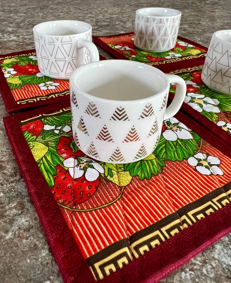 May include: White ceramic mugs with geometric patterns rest on red and green strawberry-themed placemats. The mugs have handles and are decorated with gold and black triangle and line designs. The placemats feature strawberries, flowers, and a striped pattern.