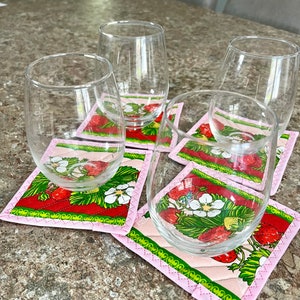 May include: A set of clear stemless wine glasses resting on square fabric coasters. The coasters feature a red background with strawberries, white flowers, and green leaves, framed by a pink border. The glasses are empty.