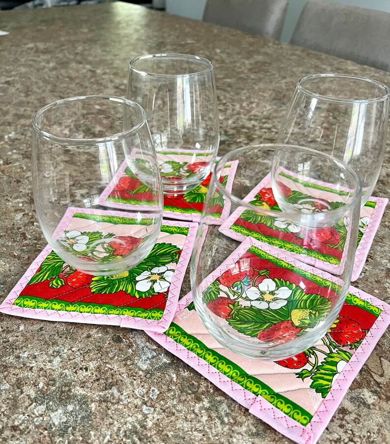 May include: A set of clear stemless wine glasses resting on square fabric coasters. The coasters feature a red background with strawberries, white flowers, and green leaves, framed by a pink border. The glasses are empty.