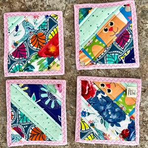 May include: Set of four square fabric coasters with a patchwork design. Each coaster features a diagonal stripe of light green fabric and colorful floral and patterned fabrics. The edges are finished with pink stitching.