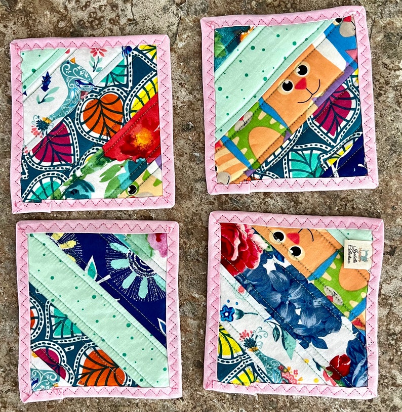 May include: Set of four square fabric coasters with a patchwork design. Each coaster features a diagonal stripe of light green fabric and colorful floral and patterned fabrics. The edges are finished with pink stitching.