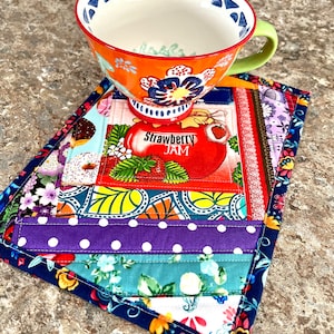 May include: A colourful patchwork fabric coaster with a blue border. The coaster features a variety of patterns, including polka dots, flowers, and geometric designs. A bright orange and blue mug with a floral design sits on top of the coaster.