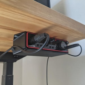 May include: A black and red audio interface, mounted under a wooden desk. The interface has multiple input jacks, knobs, and a power button. Cables are plugged into the interface. The text "Scarlett 2i2" is visible.
