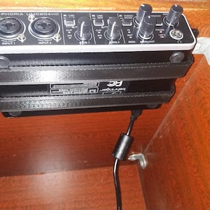Pode incluir: Uma interface de áudio preta rotulada como "U-PHORIA UMC204HD" montada dentro de um armário de madeira. A interface possui várias tomadas de entrada, botões e um cabo de alimentação. A unidade é fixada com suportes pretos e um clipe de montagem branco.