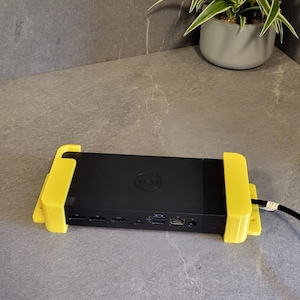 Op de afbeelding: Een zwarte Dell docking station vastgezet door een heldergele 3D-geprinte houder. De docking station heeft meerdere poorten en is verbonden met een zwarte kabel. De opstelling staat op een grijze aanrecht, met een plant in een pot op de achtergrond.