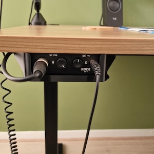 Pode incluir: Uma secretária de madeira clara com uma interface de áudio preta montada por baixo. A interface tem botões, portas e a marca "Rode". Os cabos estão ligados à interface e a um microfone. Um suporte preto suporta a secretária.