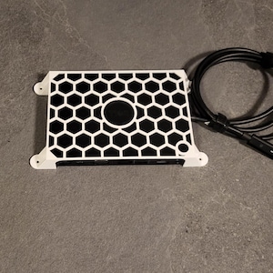 Op de afbeelding: Een witte behuizing met een zwart honingraatpatroon en een zwarte cirkel in het midden. De behuizing heeft een zwarte kabel.