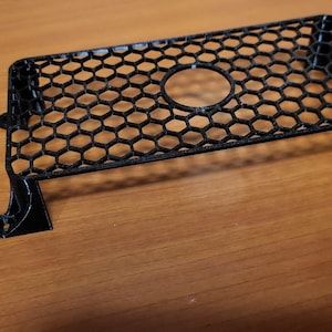 Puede incluir: Rejilla de ventilador de metal negro con un patrón de panal y una abertura circular en el centro. La rejilla tiene cuatro patas con orificios de montaje. El objeto está sobre una superficie de madera.