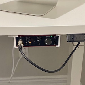 Op de afbeelding: Een zwart en rode audio-interface met knoppen en poorten is onder een witte bureau gemonteerd. Een zwarte kabel is aangesloten op de interface en een bedieningspaneel is rechts gemonteerd. Een laptop is zichtbaar op het bureau.