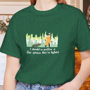 Puede incluir: Camiseta verde con un gráfico que presenta a una persona con un atuendo naranja y el texto "I should've gotten a blue spruce, they're lighter". El diseño incluye un paisaje urbano y un árbol caído. La camiseta es de material suave.