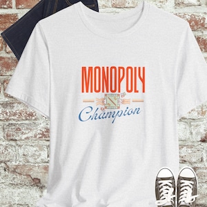 May include: White t-shirt with the text "MONOPOLY Champion" in red and blue. The shirt features a Monopoly board game graphic. Dark blue jeans and gray sneakers are also visible.