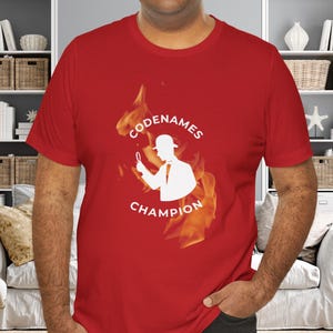 May include: Red t-shirt with a white silhouette of a person wearing a hat and holding a magnifying glass. The text "CODENAMES CHAMPION" is printed in white on the shirt, surrounded by a flame design.