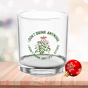 Puede incluir: Un vaso transparente con el texto "I DON'T DRINK ANYMORE, I DON'T DRINK ANY LESS, EITHER" que rodea un diseño de muérdago. Un adorno navideño rojo con un diseño de copo de nieve descansa sobre una superficie de madera.