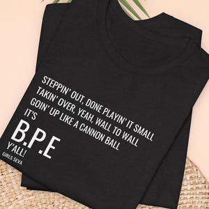 May include: Black t-shirts with white text. The text reads "STEPPIN' OUT, DONE PLAYIN' IT SMALL, TAKIN' OVER, YEAH, WALL TO WALL, GOIN' UP LIKE A CANNON BALL, IT'S B.P.E, Y'ALL! GIRLS 5EVA". The shirts are folded and stacked.