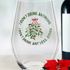 Puede incluir: Una copa de vino transparente sin tallo con el texto "I DON'T DRINK ANYMORE, I DON'T DRINK ANY LESS, EITHER" en verde. Un diseño de muérdago con bayas rojas y un lazo rojo está en el centro. Una flor de pascua roja y una botella de vino están en el fondo.