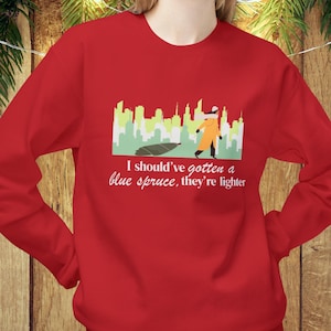 Puede incluir: Una sudadera roja con un gráfico que presenta un paisaje urbano, una persona con un traje naranja y un árbol de Navidad caído. El texto dice: "Debería haber comprado un abeto azul, son más ligeros."