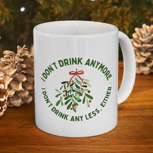 Puede incluir: Taza de cerámica blanca con el texto "I DON'T DRINK ANYMORE, I DON'T DRINK ANY LESS, EITHER" en verde, rodeando un diseño de muérdago. El muérdago tiene hojas verdes y bayas rojas, atadas con un lazo rojo. La taza está sobre una superficie de madera.