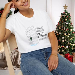 Puede incluir: Camiseta blanca con el texto "I should've gotten the blue spruce, they're lighter" y un gráfico de un edificio y un árbol. La camiseta se combina con vaqueros azules. Un árbol de Navidad decorado está en el fondo.