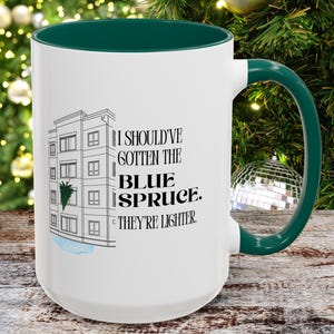 Puede incluir: Taza de cerámica blanca con interior y asa verdes. La taza presenta un gráfico de un edificio con un abeto azul y el texto "I SHOULD'VE GOTTEN THE BLUE SPRUCE. THEY'RE LIGHTER."
