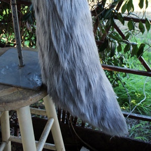 May include: A long, gray, fluffy tail made of faux fur. The tail is attached to a blue fabric base and is displayed on a white wooden stool.