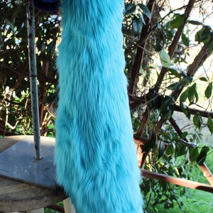May include: A long, fluffy turquoise tail made of faux fur. The tail is hanging from a stool and is against a backdrop of green foliage.
