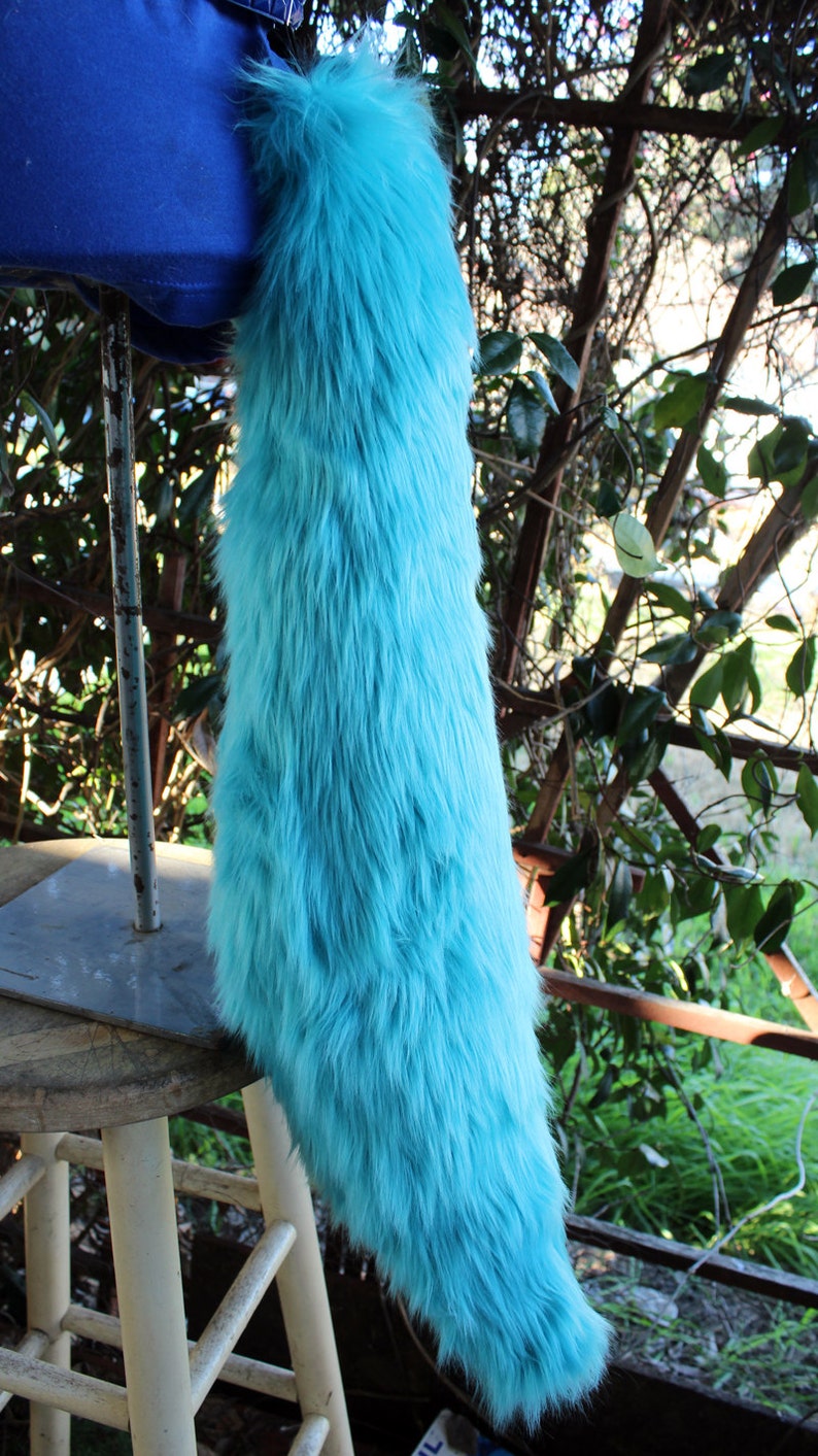May include: A long, fluffy turquoise tail made of faux fur. The tail is hanging from a stool and is against a backdrop of green foliage.