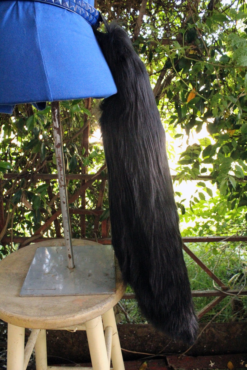 May include: A black faux fur tail with a blue fabric base and a silver metal stand. The tail is attached to the stand with a black strap. The stand is on a wooden stool.