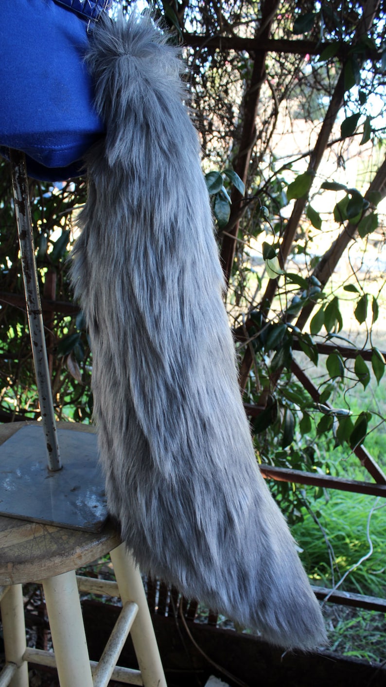 May include: A long, gray, fluffy tail made of faux fur. The tail is attached to a blue fabric base and is displayed on a white wooden stool.