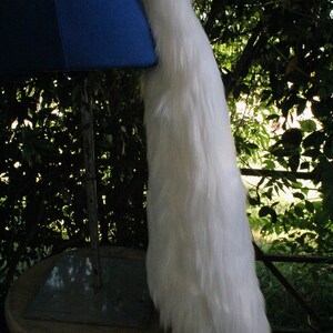 May include: A white fluffy tail prop, attached to a blue fabric base, is displayed on a wooden stool. The tail is long and full, with a soft, plush texture.